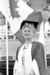 Image of Clara Thomas with cap and academic robes on standing in front of a Canadian flag, 1986. Clara Thomas fonds, F0432, image no. ASC00510.