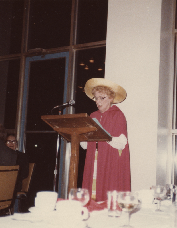 Image of Clara Thomas wearing red academic robes with a wide brimmed hat speaking at a lectern, ca. 1984. Clara Thomas fonds, F0432, image no. ASC00511.