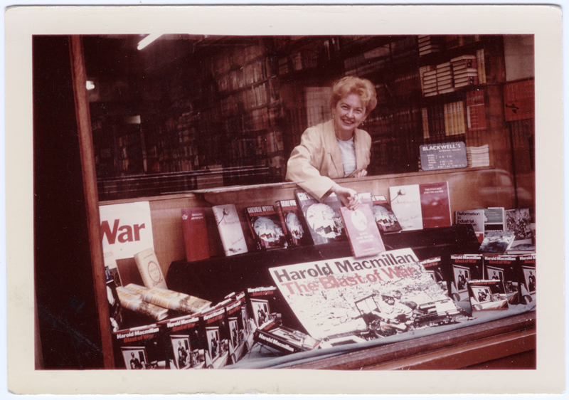 Image of Clara Thomas leaning over the display window of Blackwell's Book Store in Oxford, England, holding a copy of her book above a display featuring Harold Macmillan's "The Blast of War". Clara is smiling out to the photographer. 1967. Clara Thomas Archives & Special Collections, Clara Thomas fonds, F0432, image no. ASC07977.