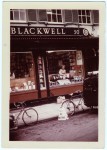Image of Clara Thomas leaning over the display window of Blackwell's Book Store in Oxford, England, holding a copy of her book above a display featuring Harold Macmillan's "The Blast of War". Clara is smiling out to the photographer, who is positioned from a distance, probably in the middle of the street, 1967. Clara Thomas Archives & Special Collections, Clara Thomas fonds, F0432, image no. ASC07978.